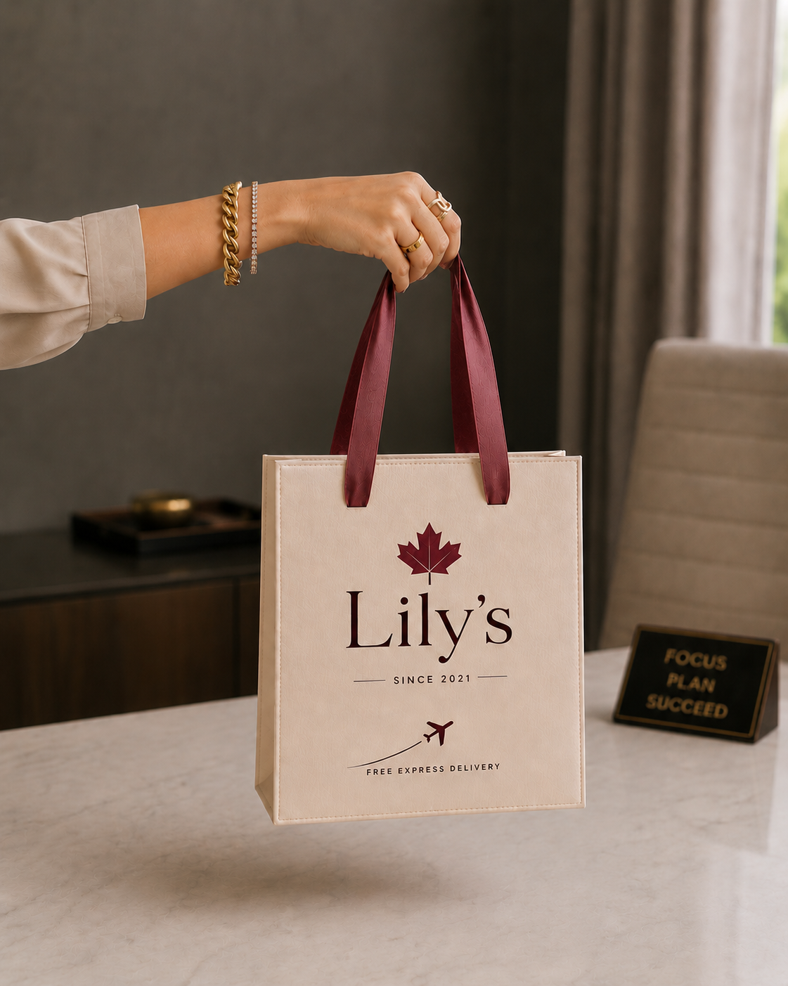 Free Express Delivery Shopping Bag featuring a cream paper gift bag with burgundy ribbon handles, Lily's logo branding, maple leaf design, and premium retail packaging presentation held by hand indoors.