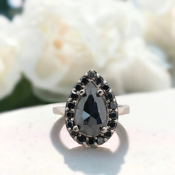 Pear-shaped black diamond halo engagement ring featuring a salt-and-pepper black diamond with galaxy-inspired inclusions, rose gold pear-cut centre stone, round black side stones, and a slim 1.80mm band.